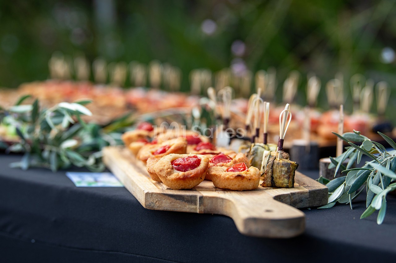 Plateau en bois avec amuse-bouches variés, dont des bouchées aux tomates et des brochettes, sur une table noire en extérieur.
