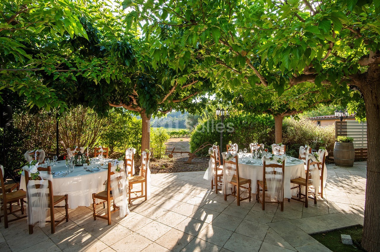 Tables rondes dressées pour un repas en extérieur, disposées sous des arbres offrant une ombre naturelle.