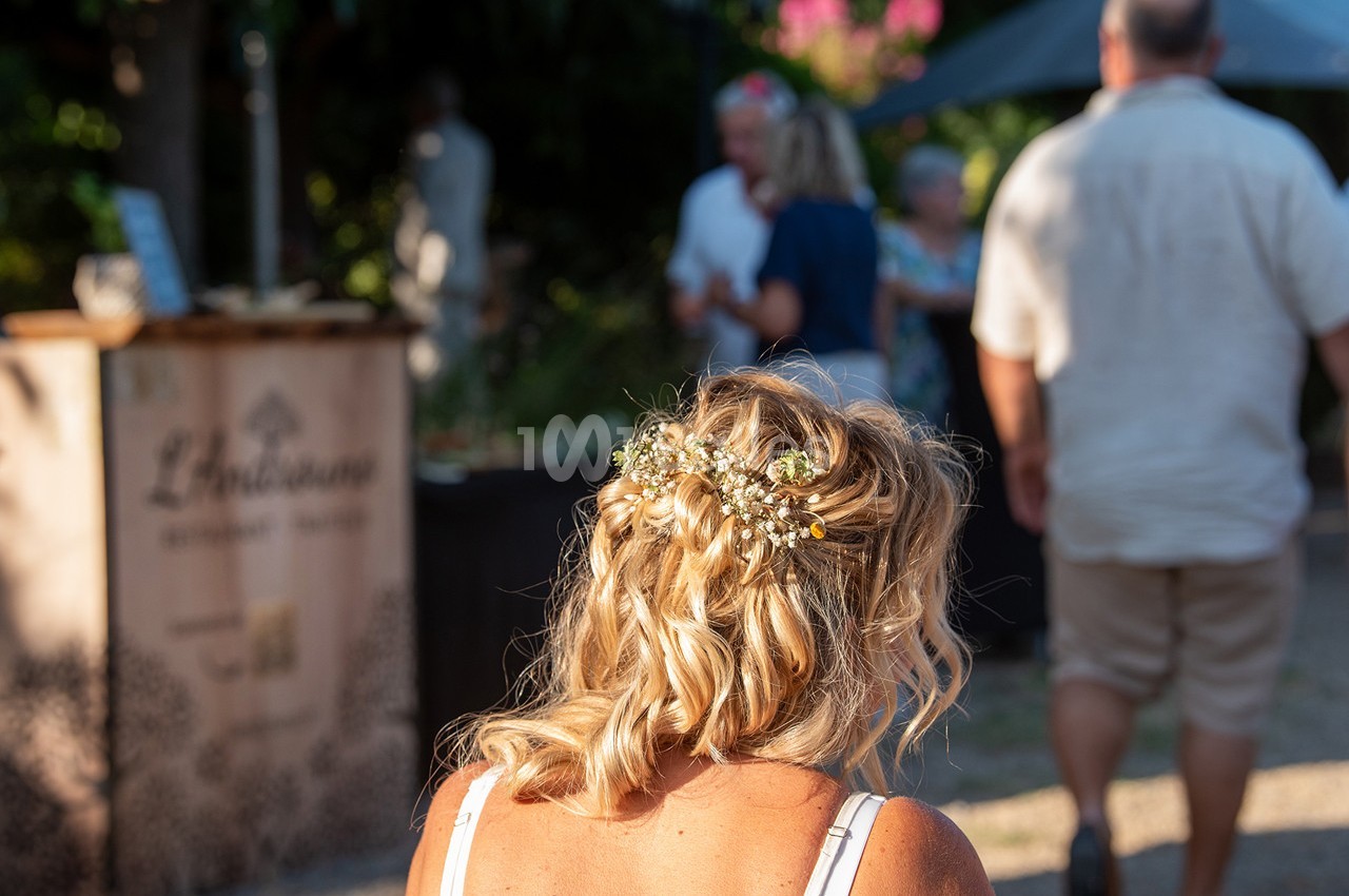 Une femme de dos avec des cheveux blonds coiffés de fleurs, dans un cadre extérieur ensoleillé avec des personnes en arrière…