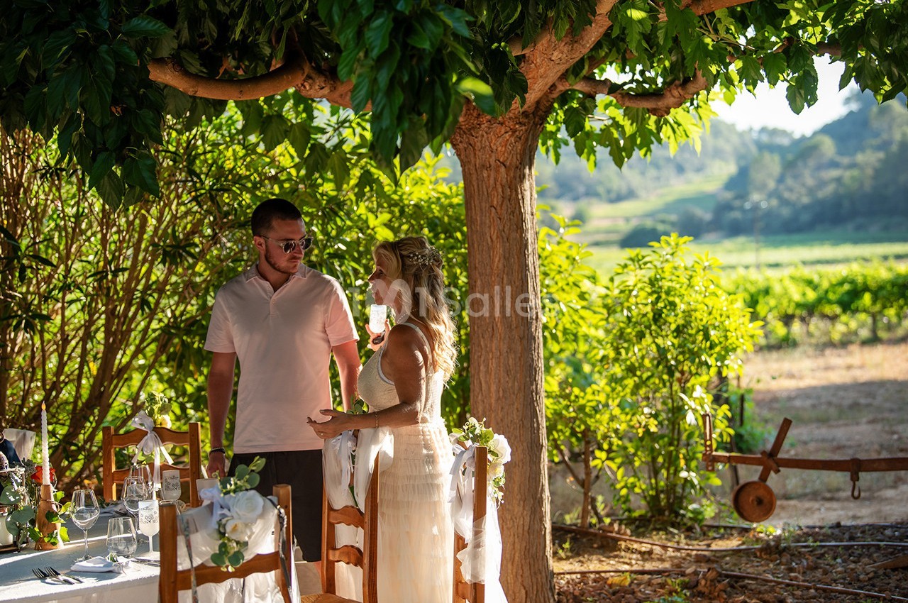 Un couple discute près d'une table décorée sous un arbre, dans un cadre champêtre et ensoleillé.