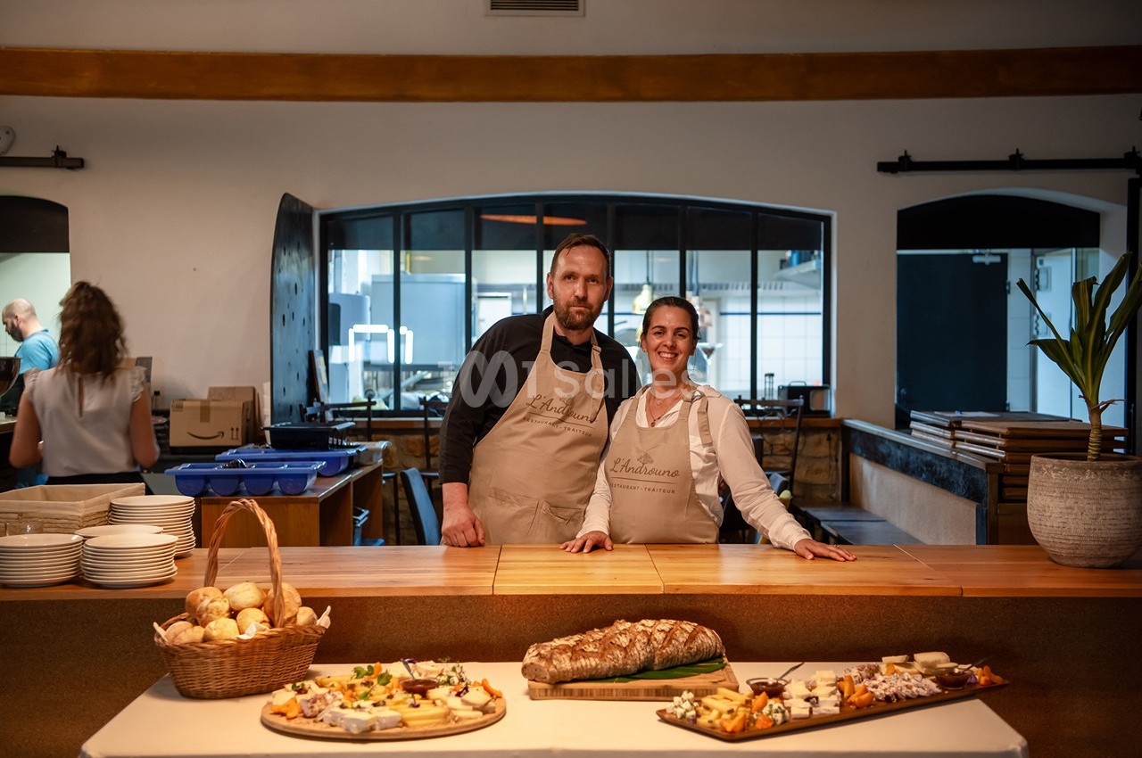 Un homme et une femme portant des tabliers se tiennent derrière un comptoir garni de pain et de plateaux de nourriture.