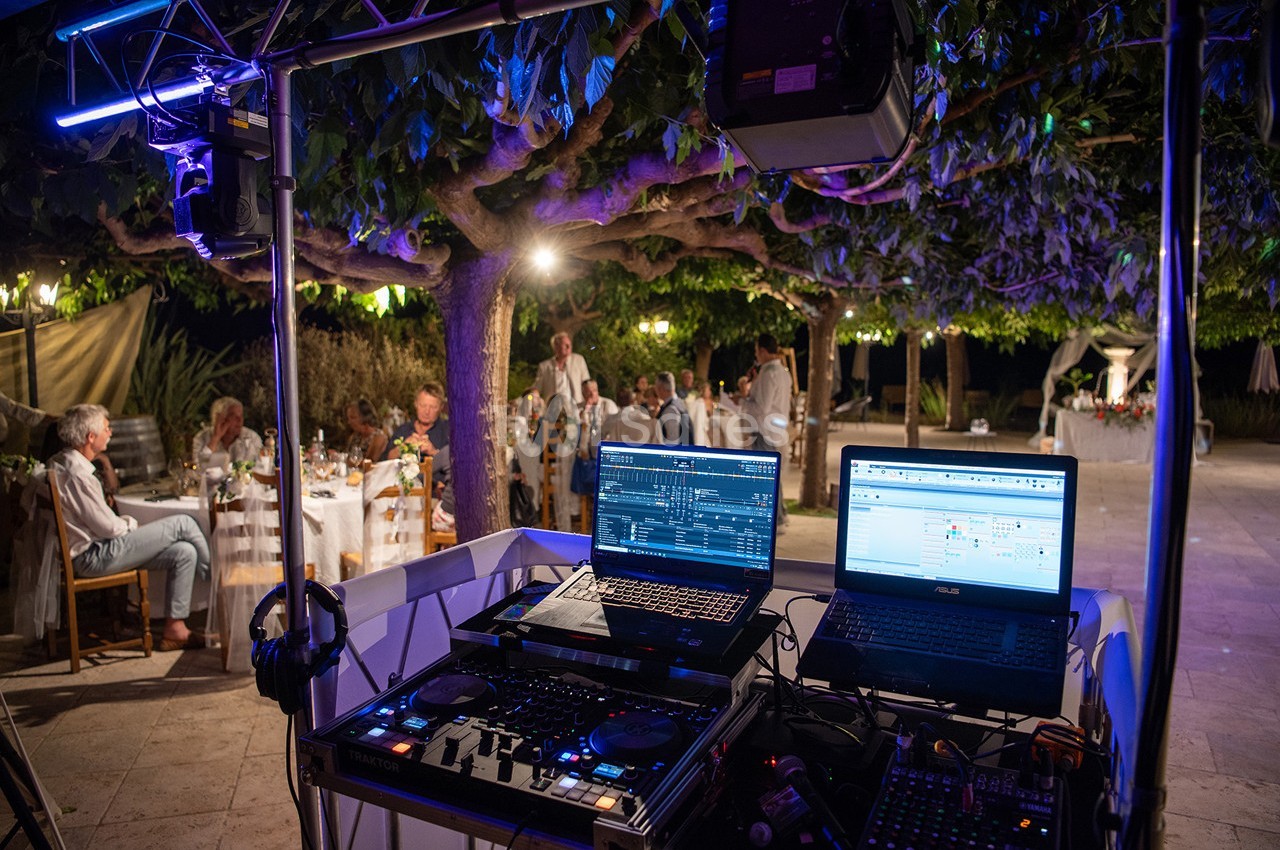 Poste de DJ avec ordinateurs et matériel audio lors d'une soirée en extérieur sous des arbres illuminés.