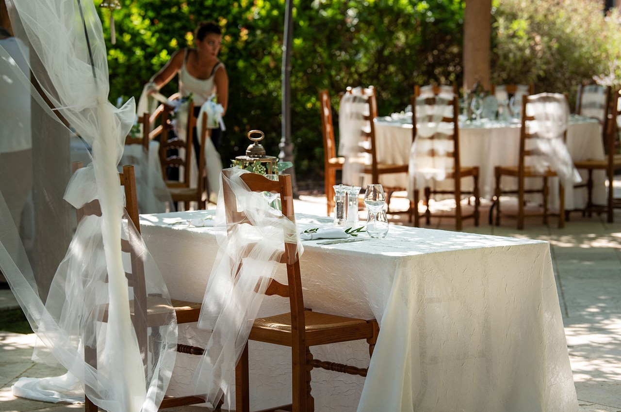 Table dressée avec une nappe blanche, entourée de chaises en bois, dans un jardin ensoleillé avec une personne en arrière…