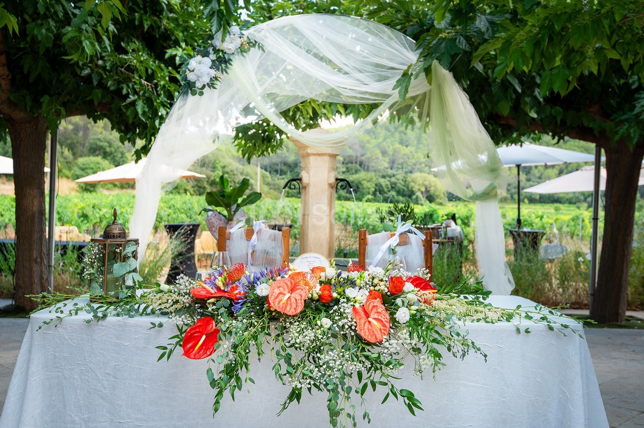 Table décorée avec des fleurs colorées et un voile blanc, installée en extérieur sous des arbres.