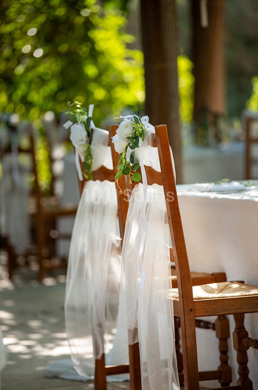 Chaises en bois décorées de rubans blancs et de fleurs blanches dans un cadre extérieur lumineux.