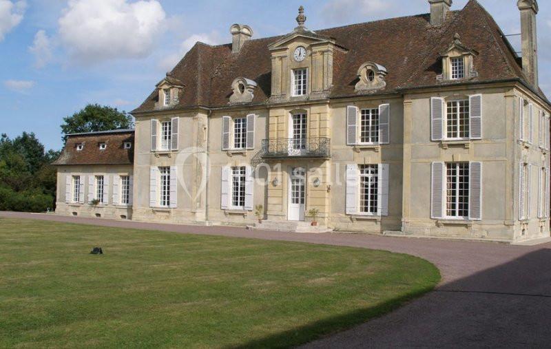 Façade d'un manoir ancien en pierre avec toit en ardoise, entouré d'une pelouse et d'un ciel partiellement nuageux.