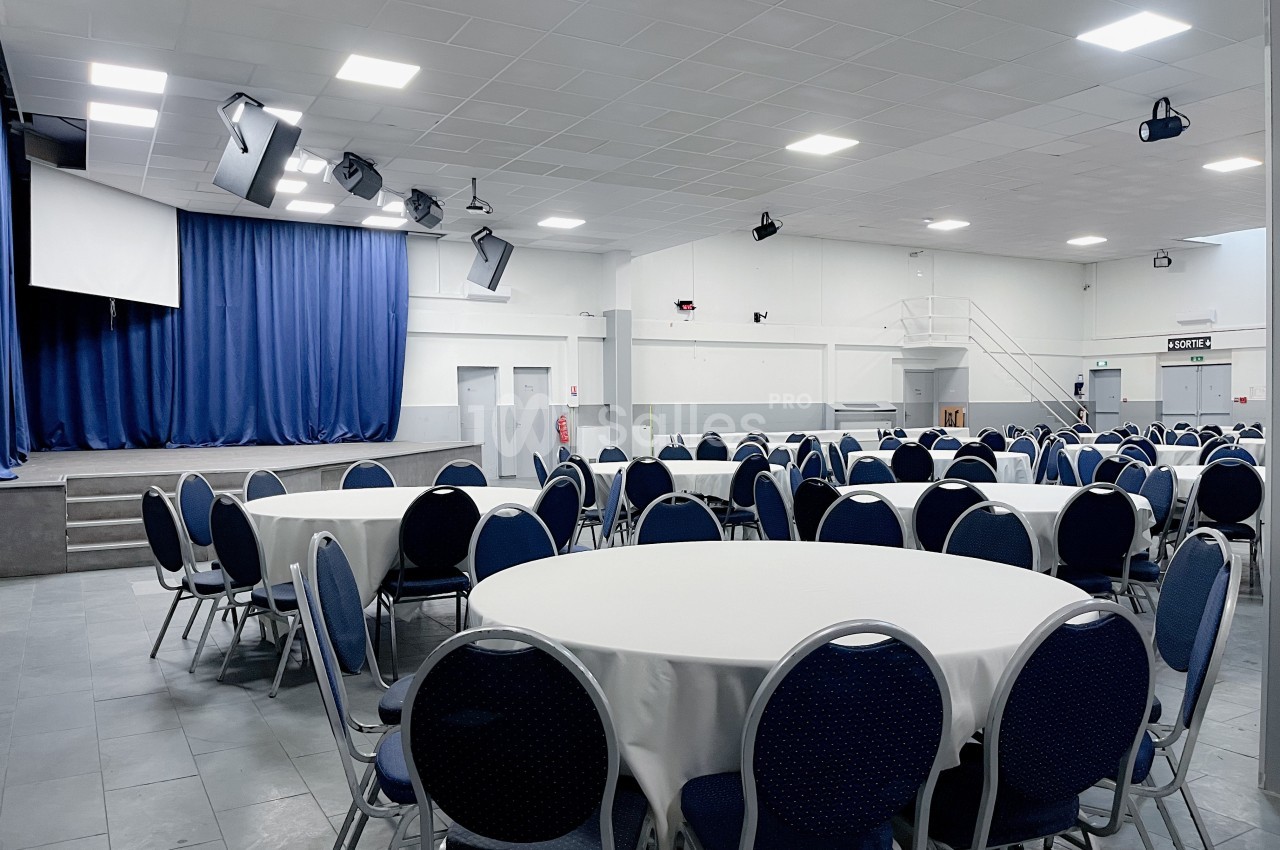 Salle de réception avec tables rondes dressées de nappes blanches, chaises bleues et une scène avec rideau bleu.