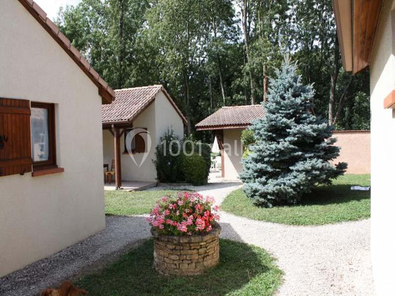Maisons avec façades claires entourées de végétation, un puits fleuri et un chemin gravillonné au premier plan.