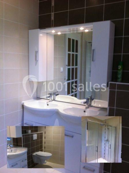 Salle de bain avec double vasque blanche, miroir éclairé, carrelage beige et marron, et toilettes visibles en arrière-plan.