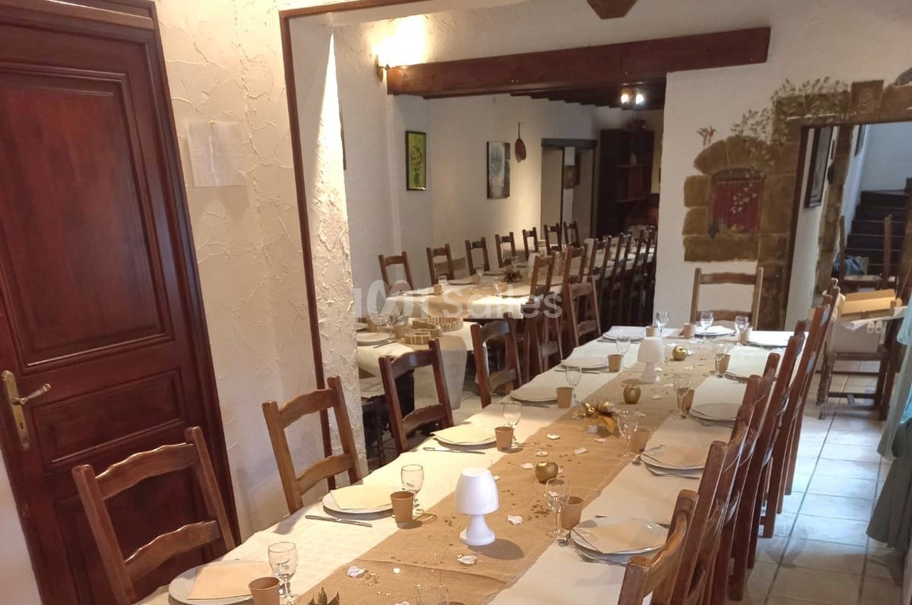 Salle de réception avec de longues tables dressées, nappes beiges, chaises en bois et décoration sobre.