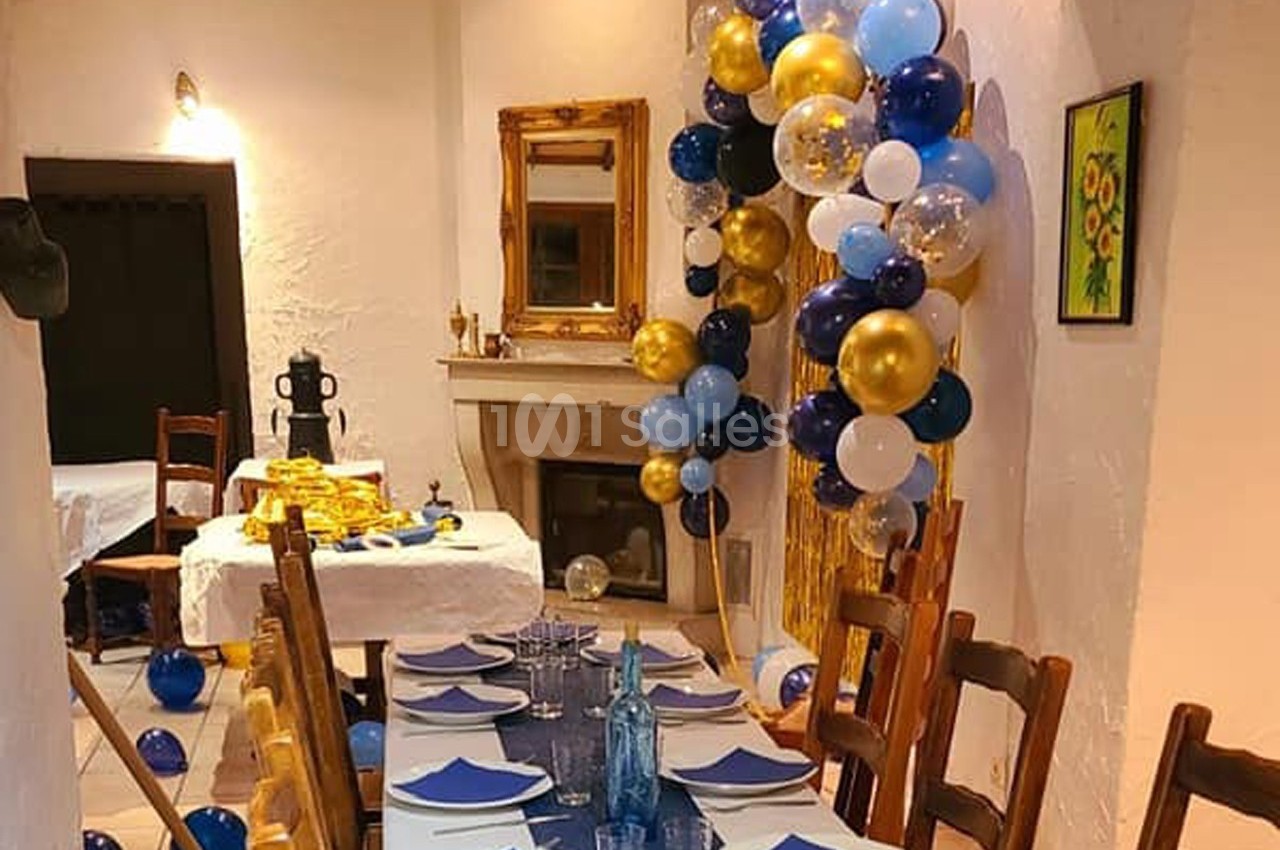 Salle décorée pour une fête avec ballons bleus, dorés et blancs, table dressée avec assiettes et serviettes bleues.