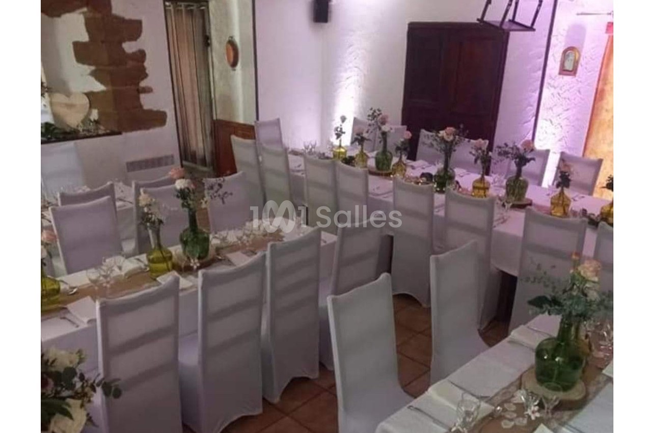 Salle décorée pour un événement avec des tables alignées, nappes blanches, chaises houssées et centres de table fleuris.