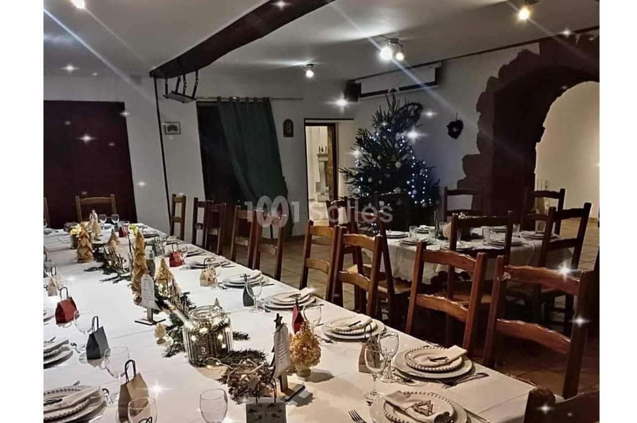 Grande salle décorée pour un repas festif, avec une longue table dressée et un sapin illuminé en arrière-plan.