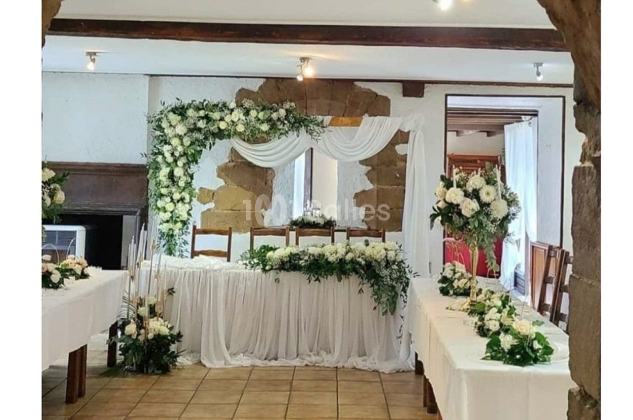 Salle décorée pour un mariage avec une table d'honneur ornée de fleurs blanches et de drapés, entourée de tables fleuries.