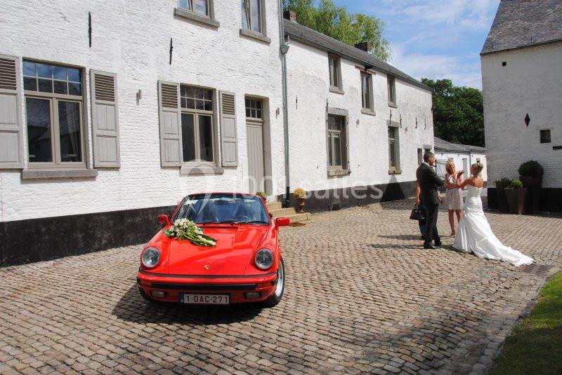 Une voiture rouge décorée pour un mariage est garée devant un bâtiment blanc, avec un couple en tenue de mariage à proximité.