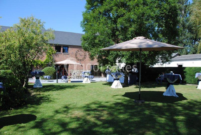 Jardin avec tables dressées pour un événement, entouré de verdure et d'un bâtiment en briques sous un ciel dégagé.