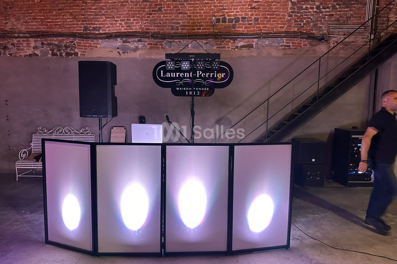 Poste de DJ avec éclairage coloré devant un mur en briques, une enseigne ’Laurent-Perrier’ et un escalier métallique.