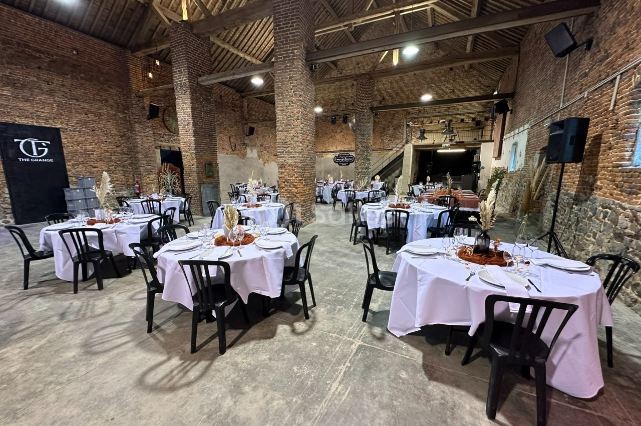 Salle en briques avec plafond en poutres apparentes, tables rondes dressées avec nappes blanches et chaises noires.