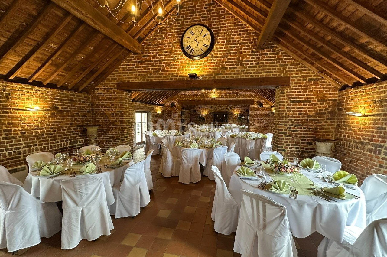 Salle de réception en briques avec tables rondes dressées, nappes blanches et serviettes pliées, sous un plafond en bois.