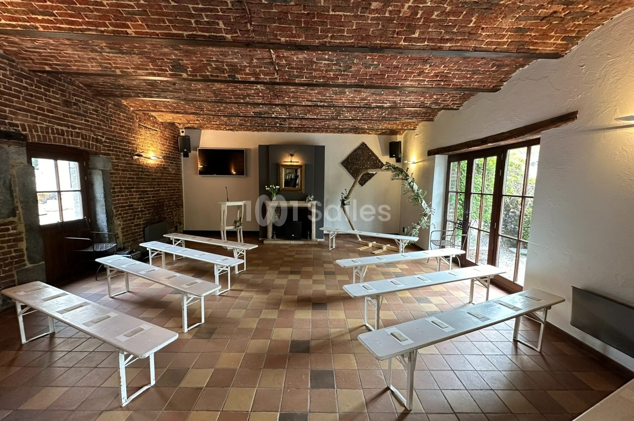 Salle en briques avec plafond voûté, bancs blancs disposés en rangées, cheminée et grande baie vitrée donnant sur un jardin.