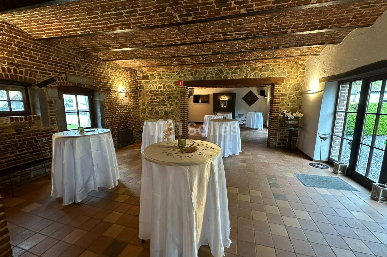 Salle en briques avec des tables hautes recouvertes de nappes blanches, éclairée par des fenêtres et des lumières murales.