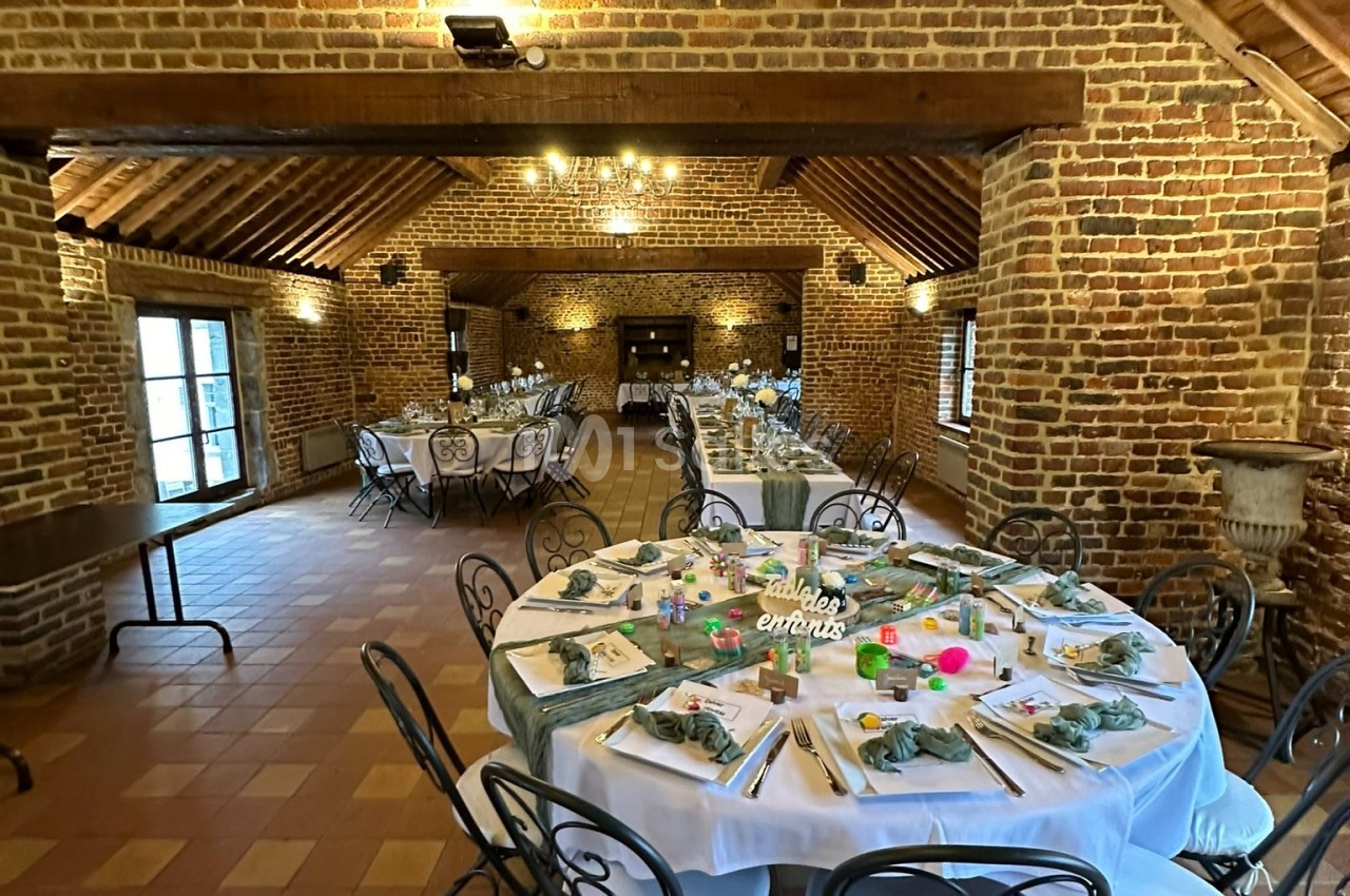 Salle en briques avec tables rondes et rectangulaires dressées pour un repas, décorées de nappes blanches et éléments…