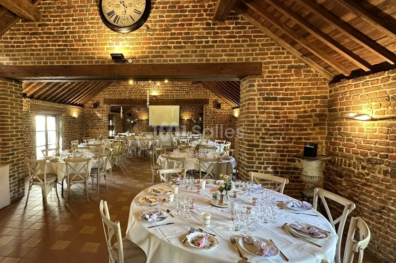 Salle de réception en briques avec tables rondes dressées, chaises en bois clair et grande horloge murale.