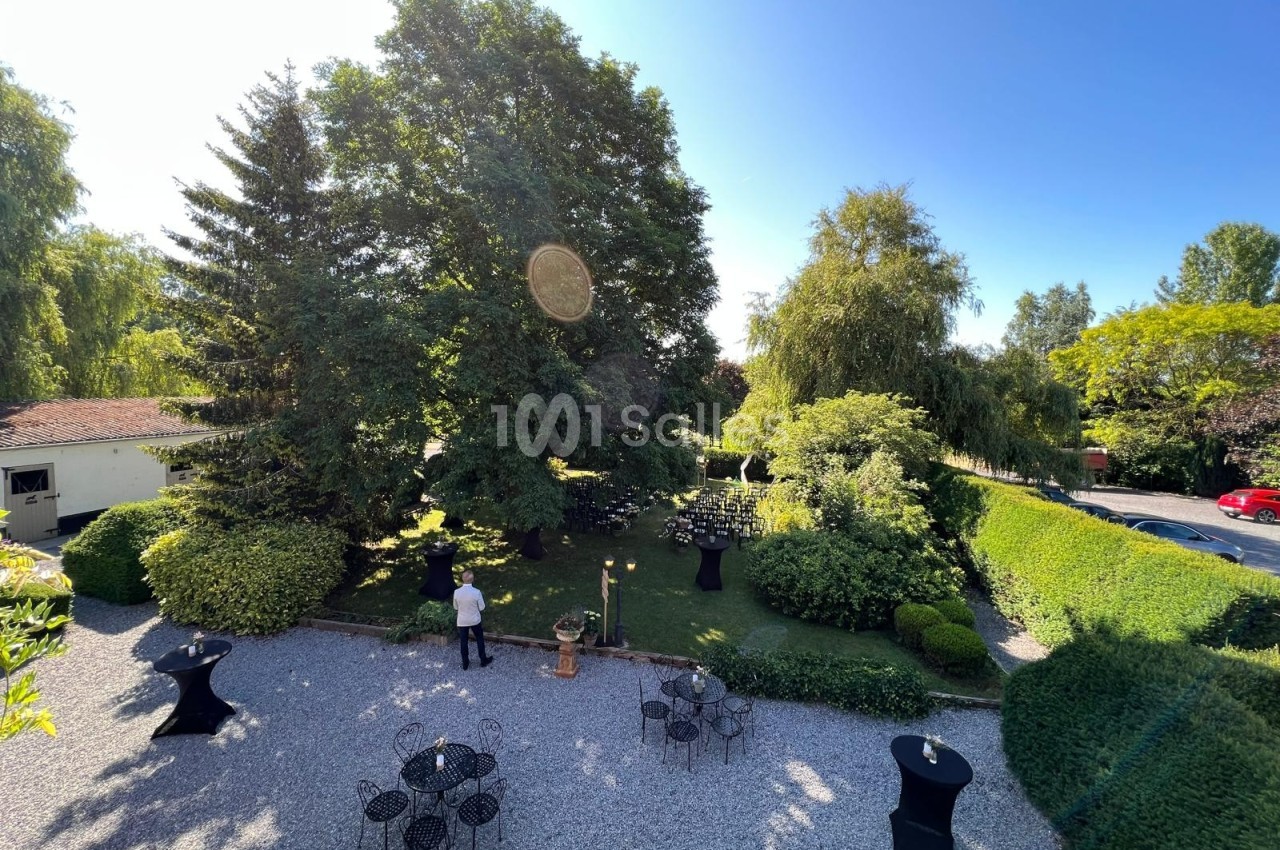Vue d'un jardin arboré avec des tables et chaises en fer forgé, un serveur debout sur une allée gravillonnée.