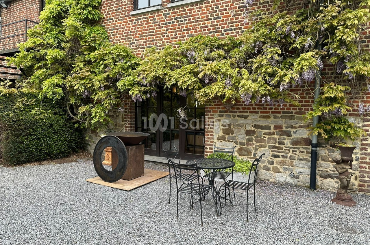 Façade en briques avec vigne grimpante, table et chaises en fer forgé sur une cour gravillonnée.