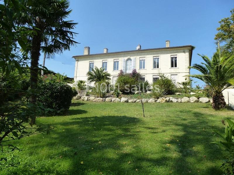 Grande maison en pierre blanche entourée d'un jardin verdoyant avec palmiers et arbustes sous un ciel bleu.