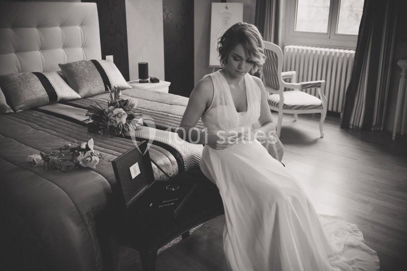 Une femme en robe blanche lit une lettre assise sur un banc dans une chambre élégante, bouquet posé sur le lit.