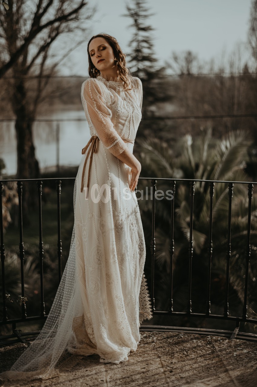Femme en robe longue en dentelle blanche posant sur un balcon avec un paysage naturel en arrière-plan.