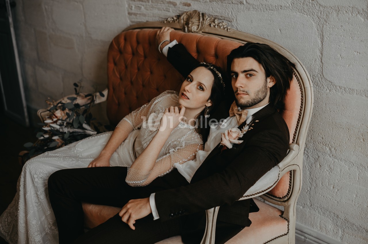 Un couple en tenue de mariage est assis sur un canapé ancien dans un décor intérieur élégant.
