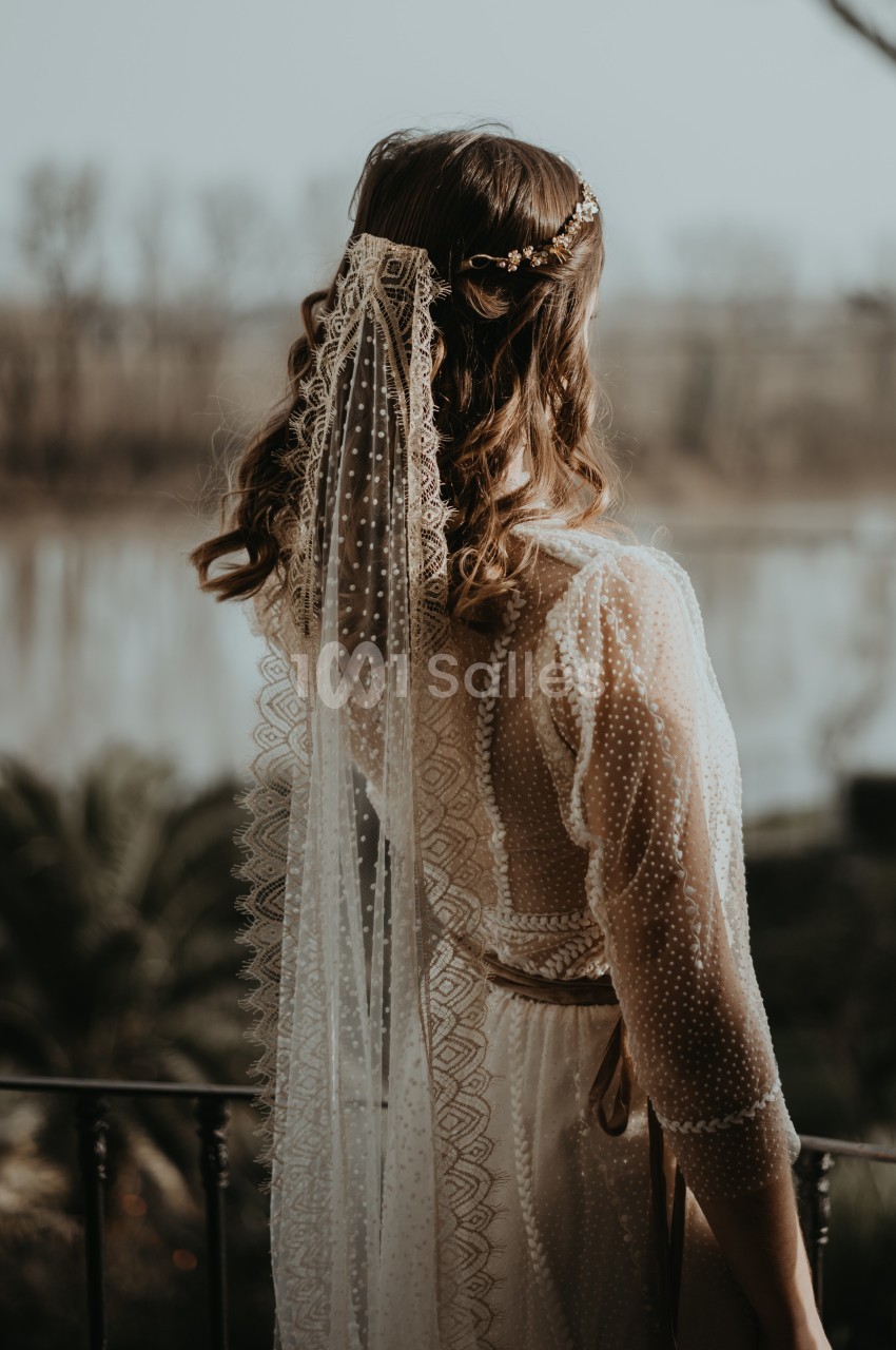 Femme de dos portant une robe en dentelle et un voile, debout sur un balcon avec un paysage fluvial en arrière-plan.
