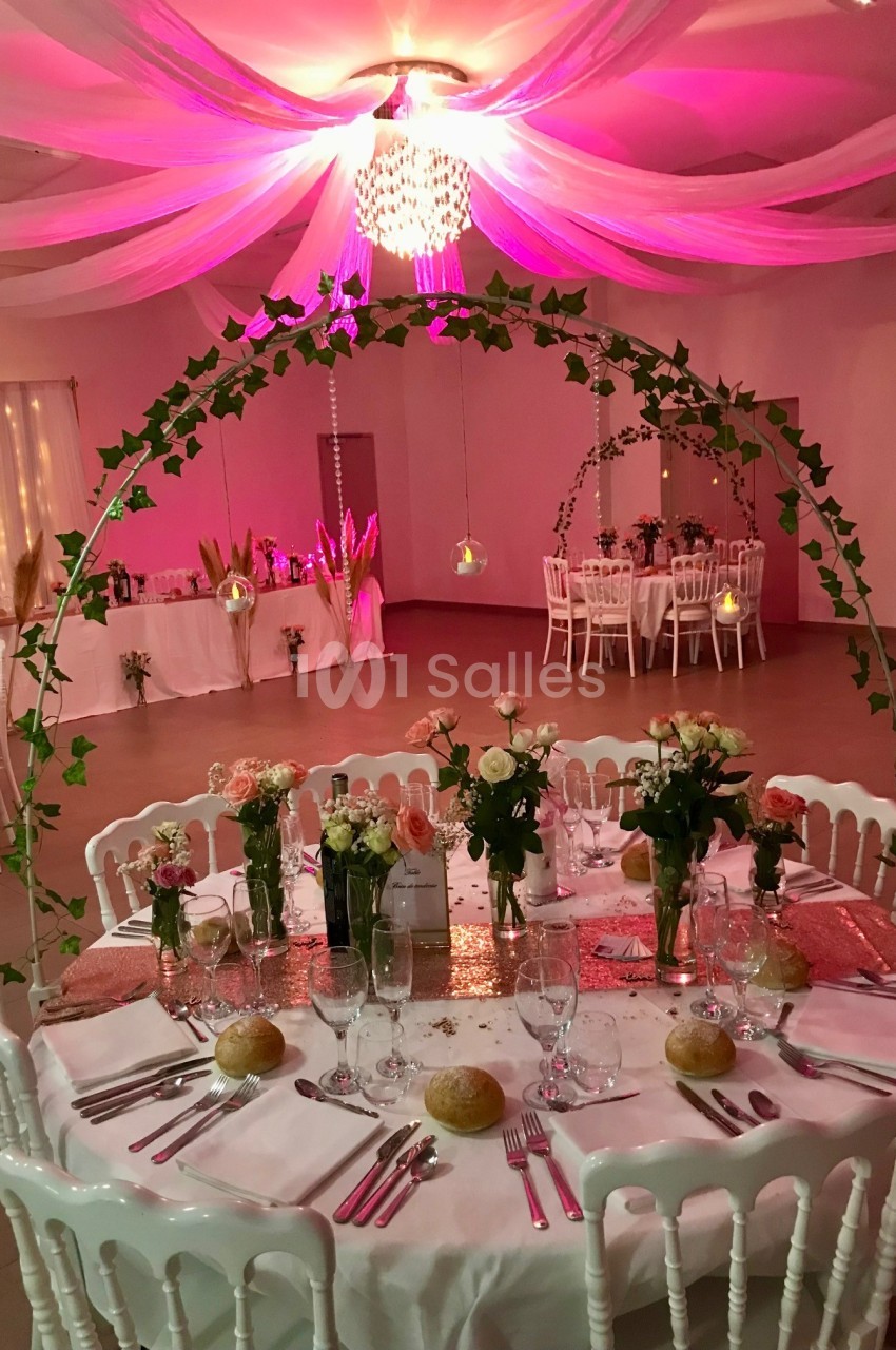 Table de réception décorée avec des fleurs, nappes blanches, et guirlandes lumineuses dans une salle éclairée en rose.
