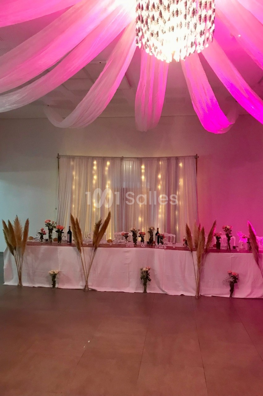 Décoration de salle avec une table longue ornée de fleurs, pampas et guirlandes lumineuses sous un éclairage rose.