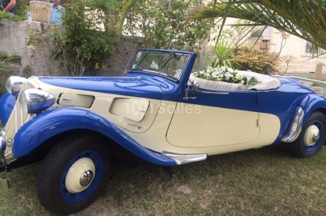 Voiture ancienne cabriolet bleu et crème, décorée de fleurs blanches, stationnée dans un jardin.