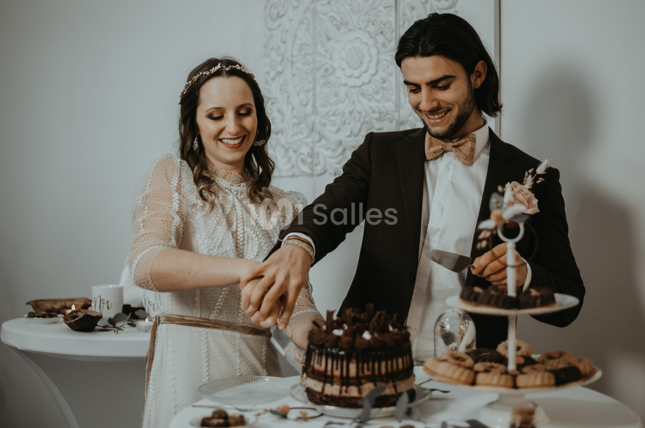 Un couple en tenue de mariage coupe ensemble un gâteau décoré de chocolat sur une table de réception.