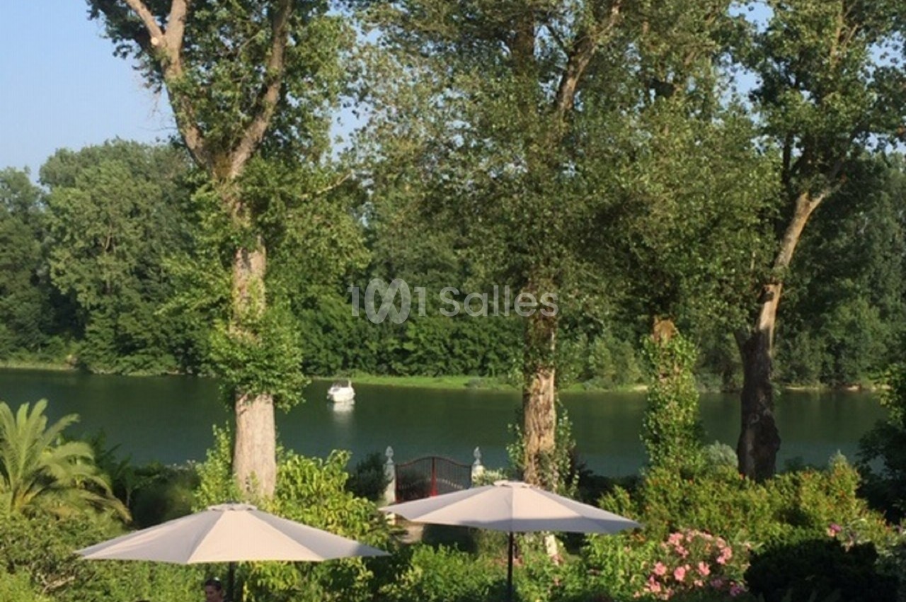 Vue sur un jardin verdoyant avec parasols, bordé par une rivière calme et entouré d'arbres en été.