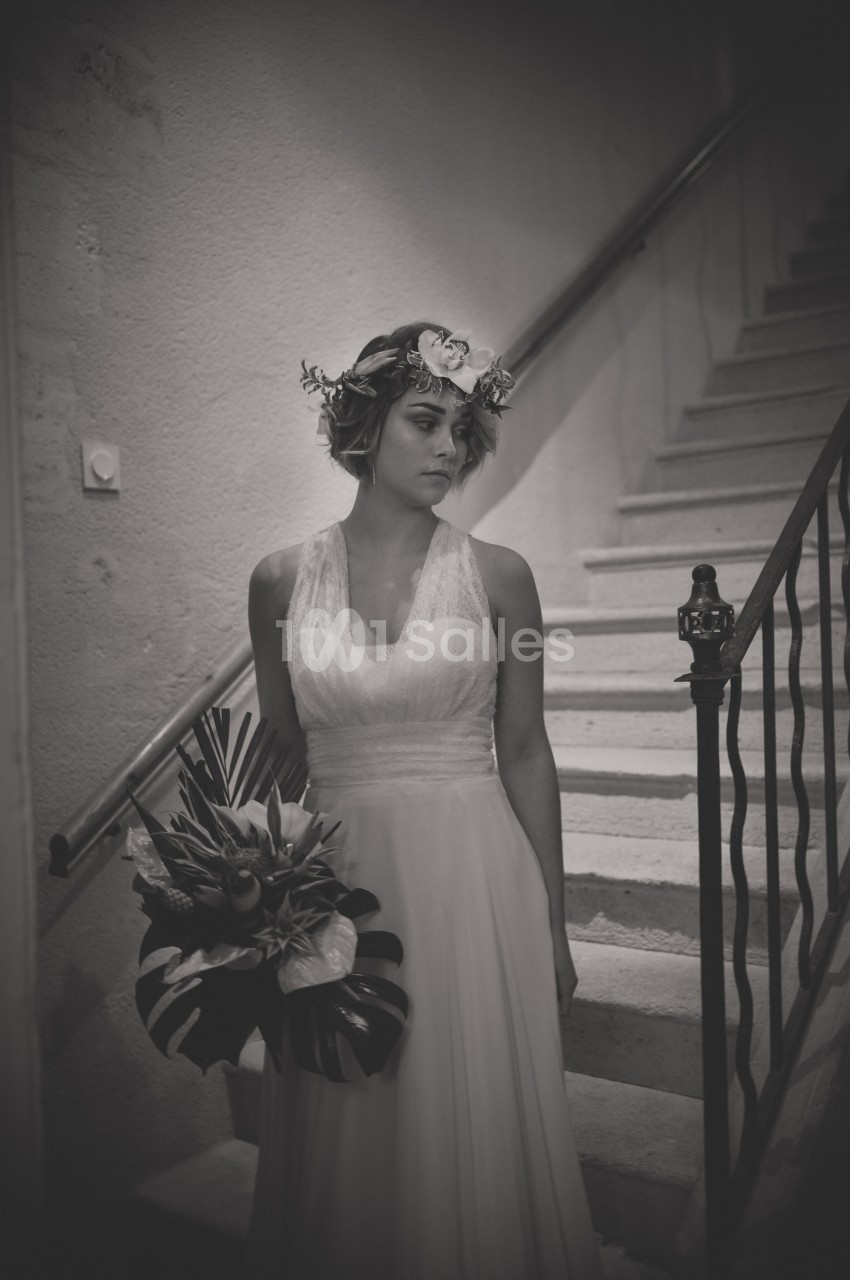 Une femme en robe de mariée tient un bouquet tropical, debout près d'un escalier en intérieur.