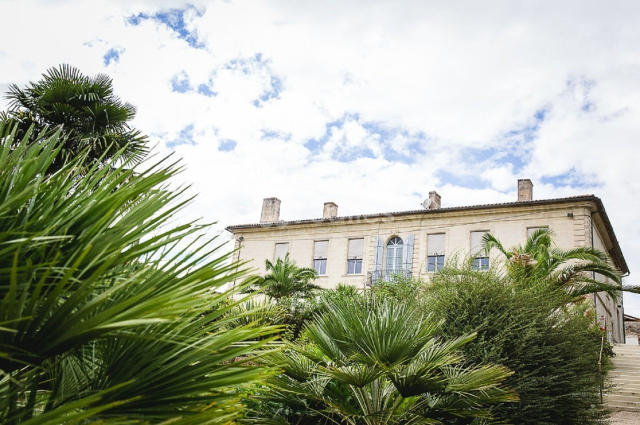 Façade d'un bâtiment ancien entouré de végétation luxuriante avec un ciel partiellement nuageux en arrière-plan.