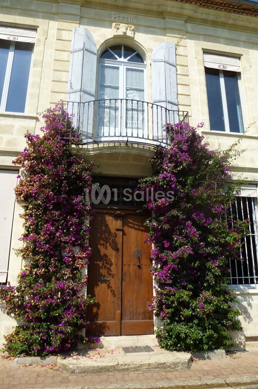 Façade en pierre avec une porte en bois encadrée de bougainvilliers fleuris et un balcon avec volets ouverts.