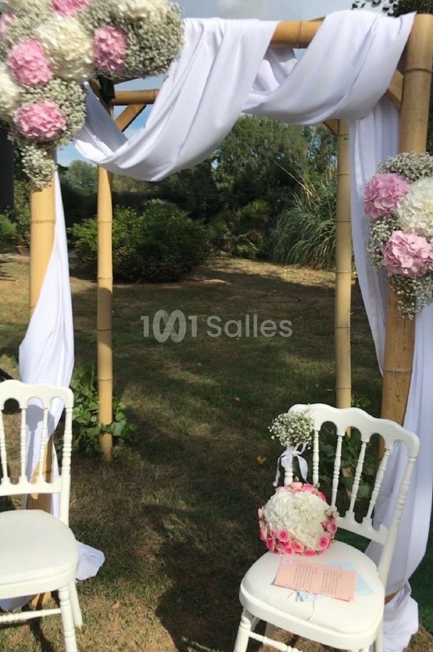 Arche de mariage en bambou décorée de fleurs roses et blanches, entourée de chaises blanches dans un jardin.