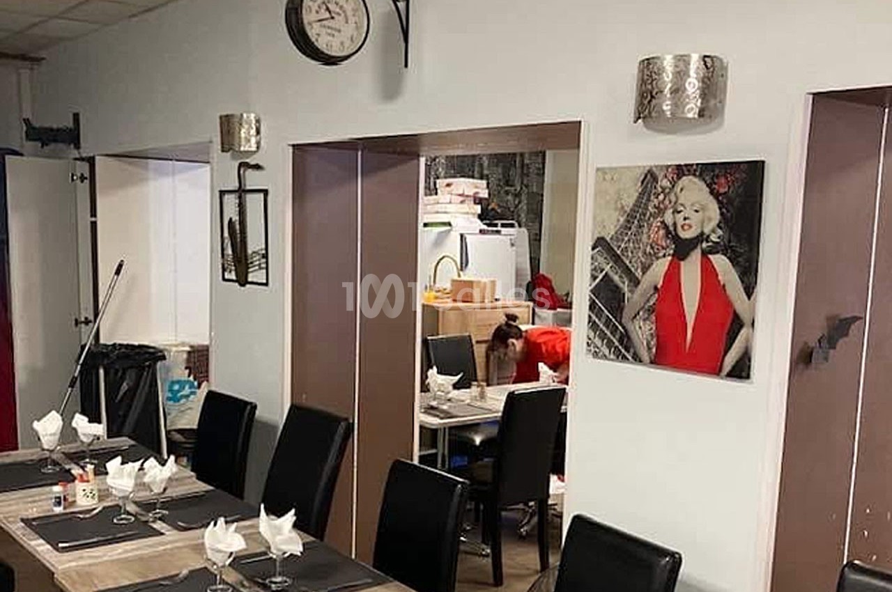 Salle de restaurant avec tables dressées, décoration murale et aperçu d'une cuisine en arrière-plan.