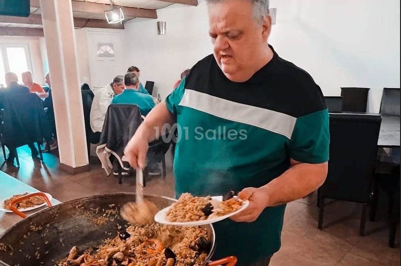 Un homme sert de la paella avec une louche dans une assiette, dans une salle avec des convives assis.