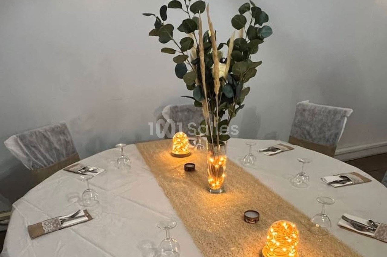 Table ronde décorée avec une nappe blanche, un chemin de table doré, des bougies lumineuses et un vase de feuillage.