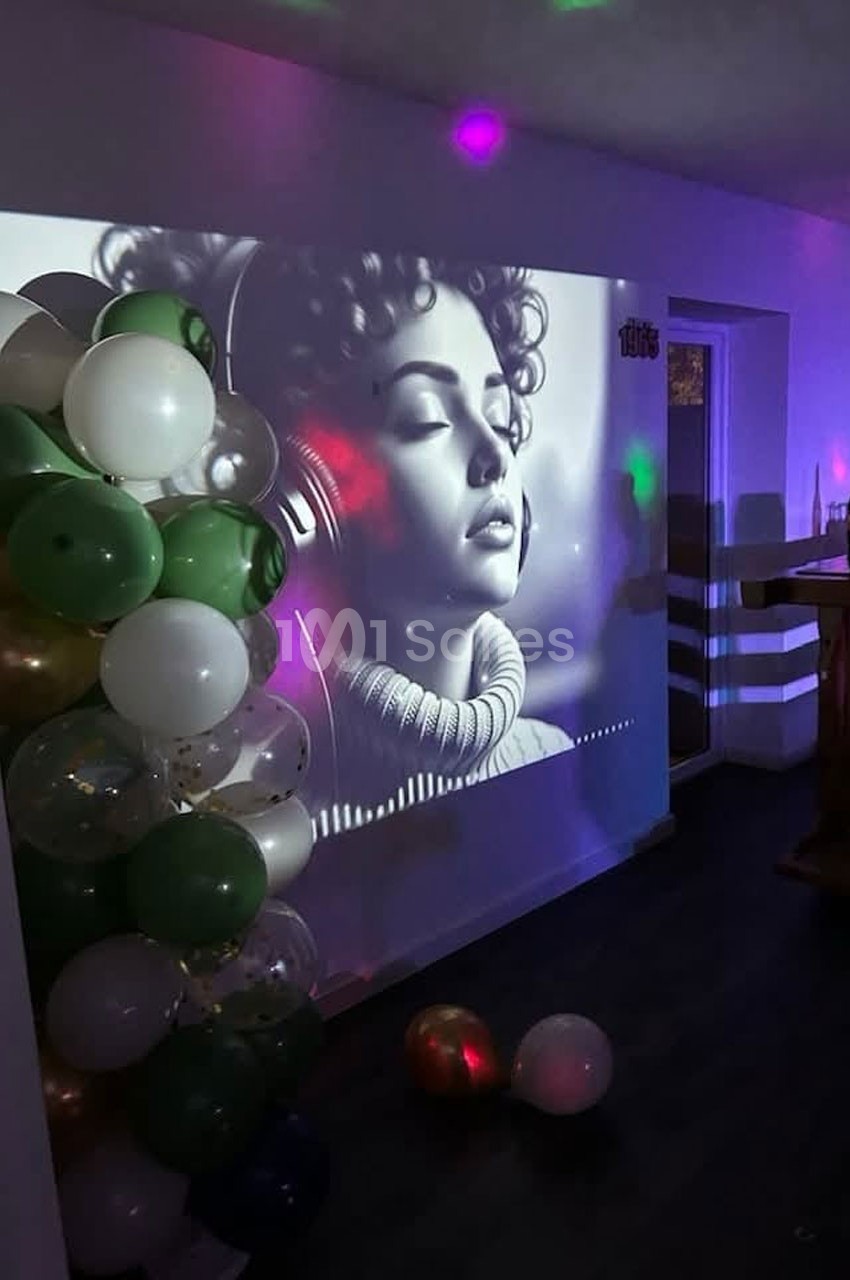 Projection en noir et blanc d'une femme avec un casque audio, entourée de ballons dans une pièce faiblement éclairée.