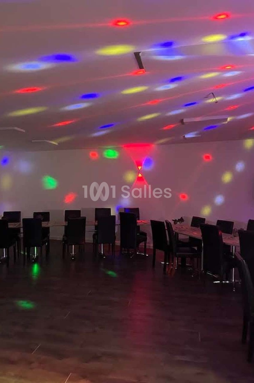 Salle sombre avec des tables et des chaises alignées, éclairée par des lumières colorées projetées au plafond.