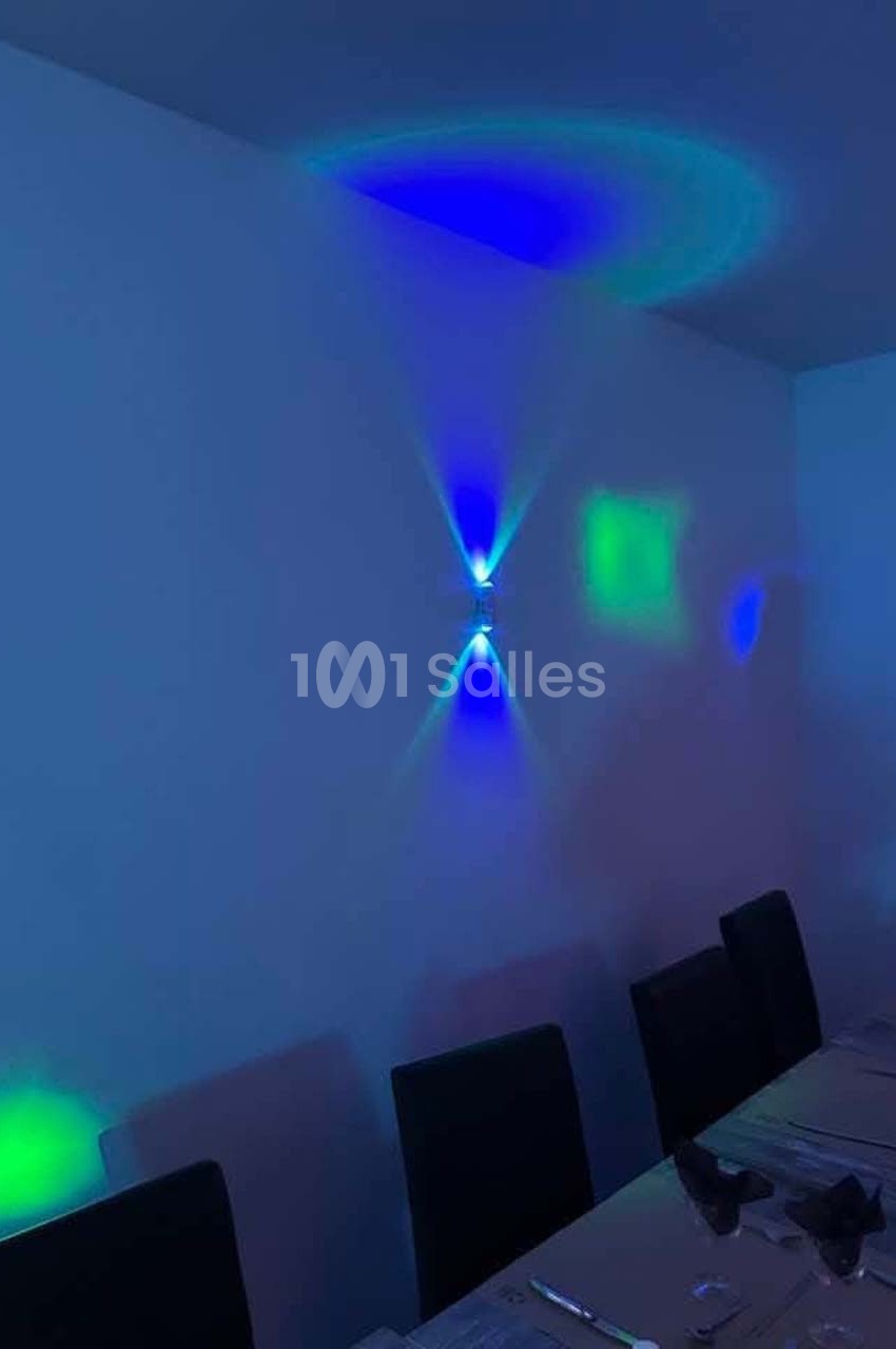 Éclairage mural diffusant des lumières bleues et vertes dans une salle avec une table dressée et des chaises noires.