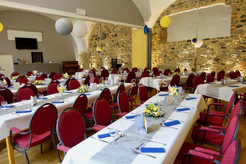Salle de réception décorée avec des tables dressées, nappes blanches, serviettes bleues et chaises rouges.